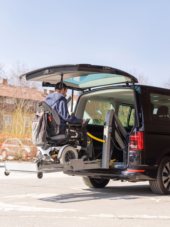 Ein Rollstuhlgerechtes Fahrzeug mit einer Hebebühne auf der eine Person im Elektrorollstuhl Von hinten in das Fahrzeug verladen wird.