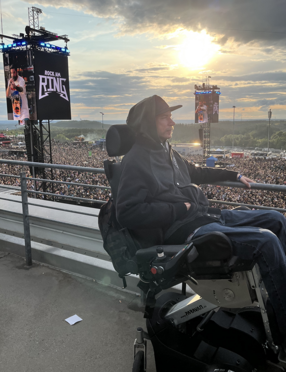 Im Hintergrund bricht die Sonne durch die Wolken. Ein E Rollstuhlfahrer steht erhöht. Im Hintergrund eine Menschenmenge auf einem Festival